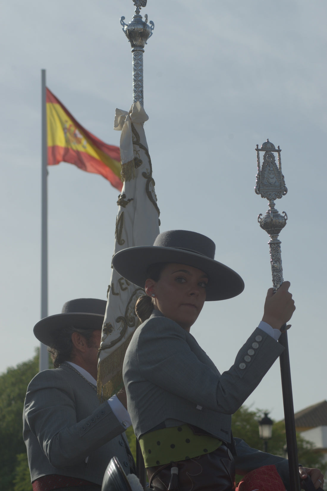 Two people in traditional Spanish attire holding a banner and staff with a flag